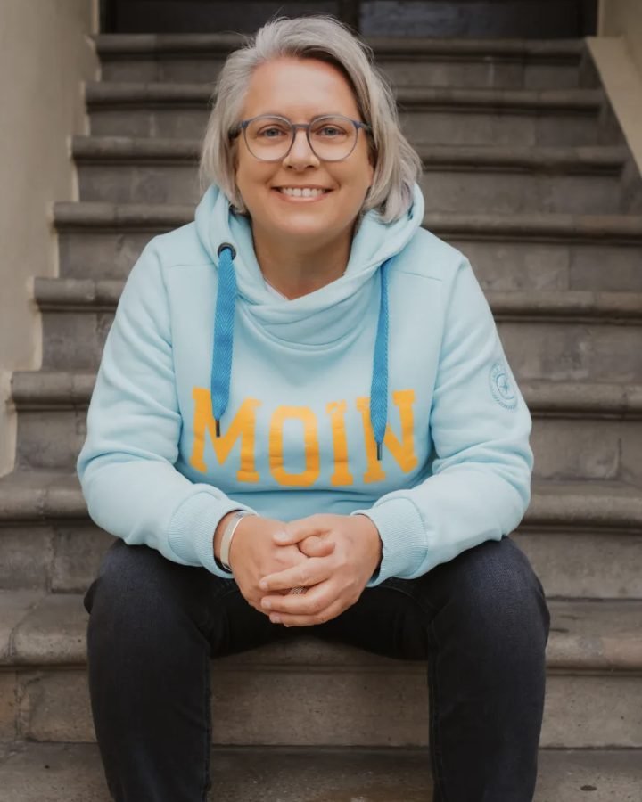 Kirsten Biema lächelnd auf einer Steintreppe, hellblauer Hoodie mit „Moin“-Schriftzug